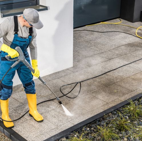 Worker in protective gear using a pressure washer to clean an outdoor patio area with a high-pressure water stream